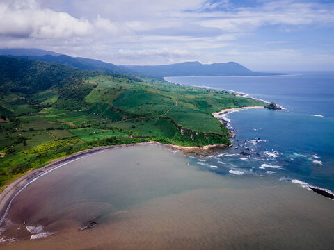 Aerial view of the lush green coastline featuring rolling hills, agricultural fields, and a sandy beach meeting the deep blue ocean in Sumbawa, Indonesia.