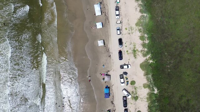 Vehicles form distinct row along sandy edge, while colorful sunshades cluster near rolling surf line. Swimmers enjoy cooling water as waves wash onto wet shoreline, Surfside Beach, Freeport, TX