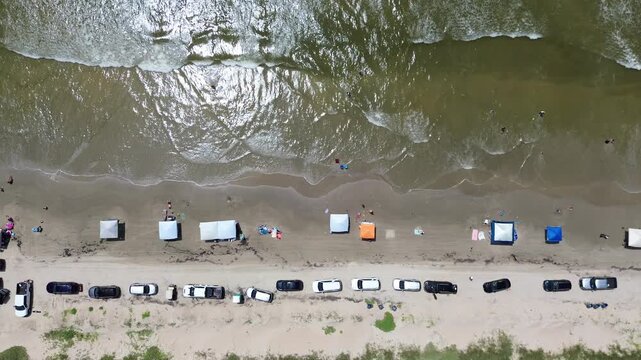 Motorists maintain linear path along dunes while beachgoers enjoy shaded structures by surf. Sunlight glints off tidal foam patterns meeting sand amidst summer heat, Surfside Beach, Freeport, TX