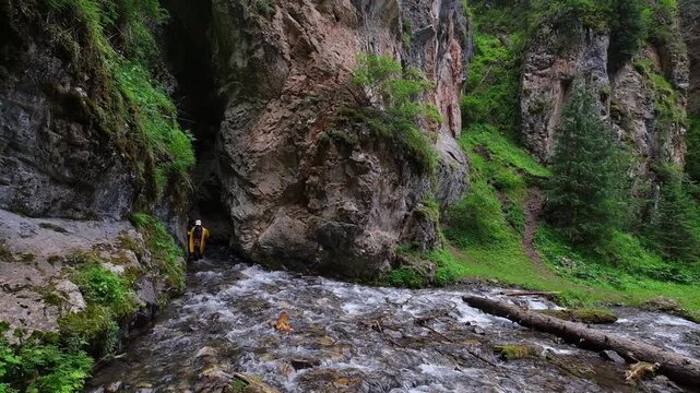 A female traveler in a yellow raincoat walking out of the narrow Tamgaly Tas gorge by a rushing river in Kolsai Lakes National Park, Kazakhstan. Commercial travel background with copy space.