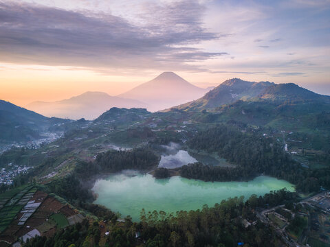 Aerial view of Telaga Warna and Telaga Pengilon lakes with lush hills, misty forests, and distant volcanic peaks under a soft sunrise sky in Kecamatan Batur, Jawa Tengah, Indonesia.