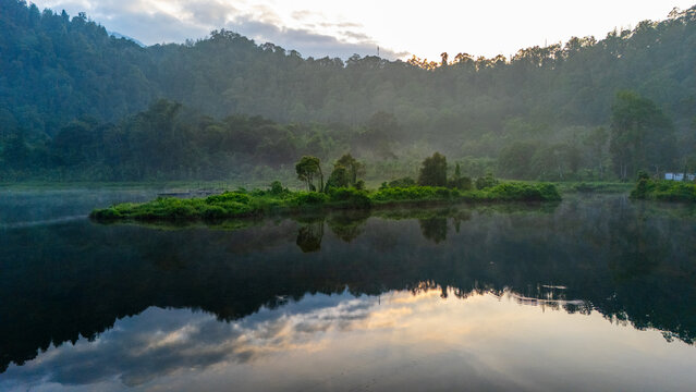 Aerial view of a serene lake with a small lush island surrounded by dense forest-covered hills under soft morning light in West Java, Indonesia.