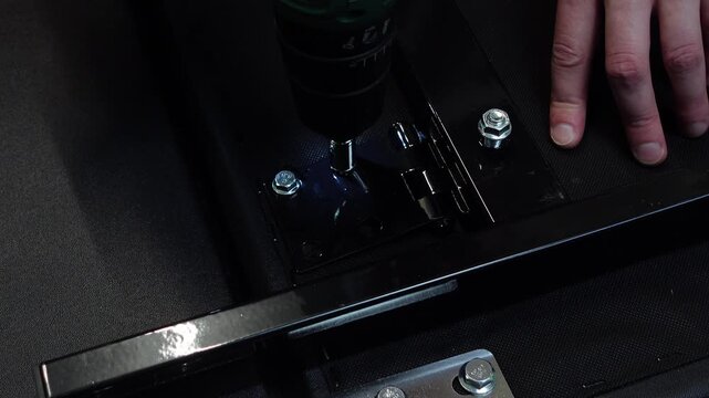 Hands using a cordless drill with a socket bit to tighten bolts into a black metal hinge during furniture assembly