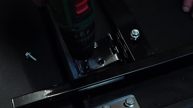 Hands using a cordless drill with a socket bit to tighten bolts into a black metal hinge during furniture assembly