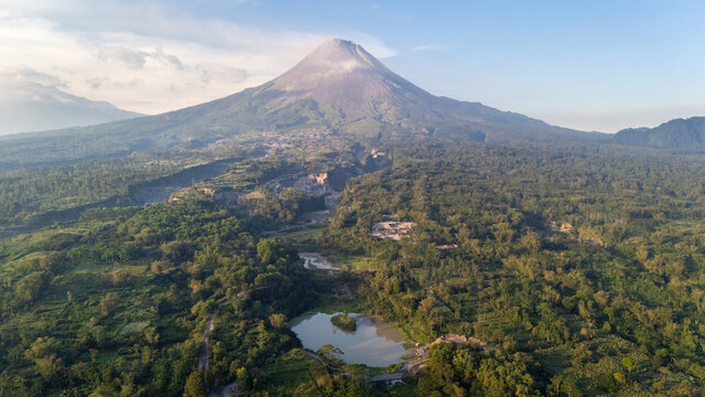 Aerial view of Mount Merapi volcano rising above lush tropical forests and a small lake in the volcanic landscape of Indonesia.