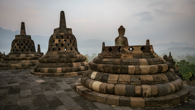 Historic stone stupas and a seated Buddha statue at Borobudur Temple in Central Java, Indonesia, overlooking a misty landscape at dawn. 