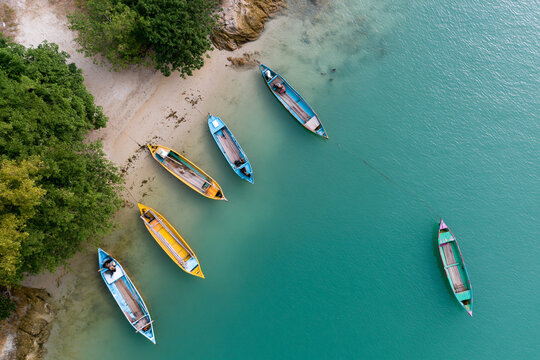 Aerial view shows colorful boats resting near a sandy shore. Turquoise water gently laps the shoreline, reflecting sky. Trees frame the left side, adding green contrast to scene