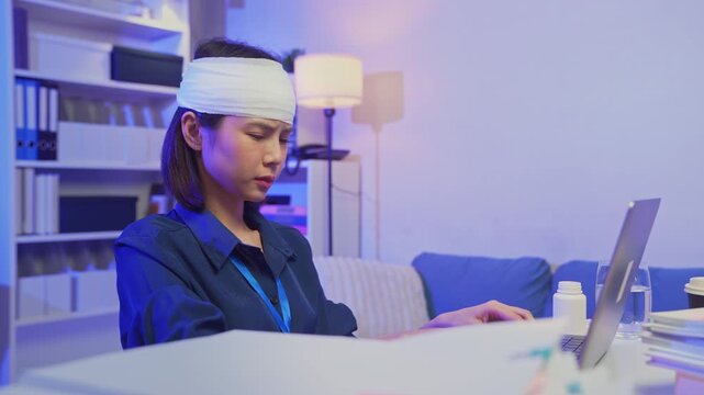 Injured young Asian office worker wearing head bandage while working on laptop in office, health and work pressure concept