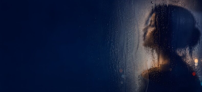 Silhouette of young woman looking through wet rainy window glass at night, moody conceptual portrait