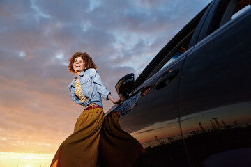 Obraz premium Young woman with curly hair leans from a car door at sunset, smiling as warm light highlights her denim jacket and flowing mustard skirt.