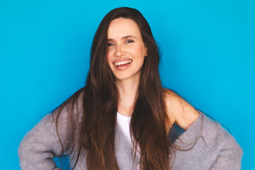Fototapeta premium Portrait of a happy young woman with long brown hair smiling genuinely at the camera, wearing a cozy oversized grey mohair sweater, isolated on a bright blue background.