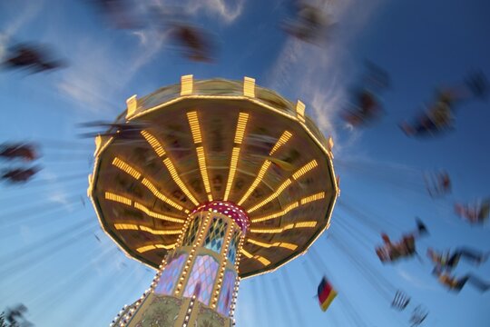 An illuminated carousel at dusk, spinning fast, in front of a blue sky with clouds and a Germany flag, Killiani Volksfest, W&uuml;rzburg, Bavaria