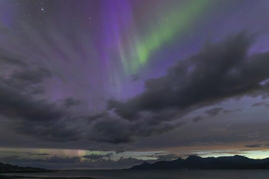 The Vestfjord in Nordland, Norway, has an enchanting atmosphere at night. Dark clouds and vivid aurora, polar lights dance over the fjord. The rugged cliffs and majestic mountains are reflected in the calm water and create a mystical backdrop