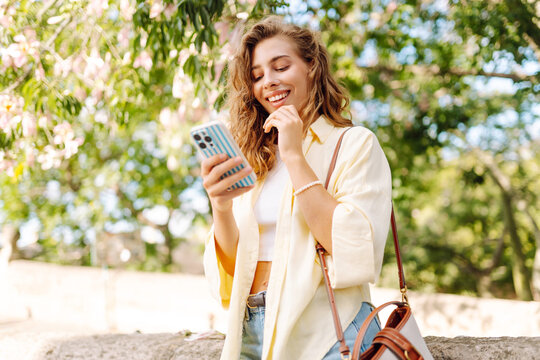 Woman stands in park with a phone in hand on sunny day. Beautiful woman blogs or texts on phone and enjoys stroll through city. Concepts of relaxation, weekends, and enjoyment.