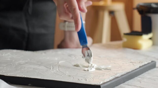Hand creates textured artwork on canvas at home spreading white plaster paste with a palette knife at a covered work table. Concept of handmade wall decor and creative hobby.