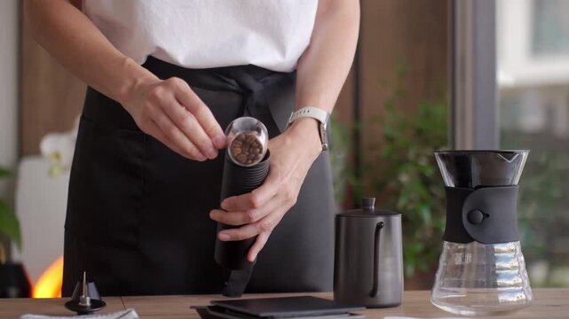 Person pouring coffee beans into hand grinder preparing coffee with kettle and pour over dripper on a wooden table. Concept of home coffee brewing.