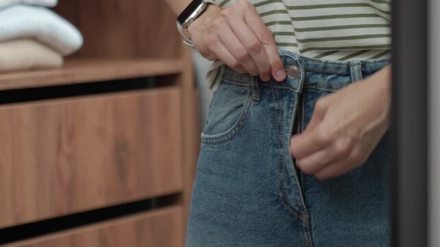 Hands fastening button on blue jeans while getting dressed in a wardrobe. Concept of denim outfit everyday fashion and personal style.