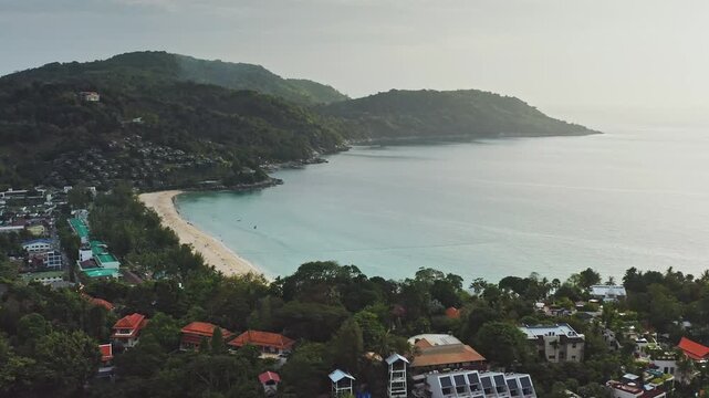 Coastal landscape sandy Kata Noi Beach meeting the clear turquoise Andaman Sea, surrounded by lush green hills, tropical vegetation and various resort buildings on a hazy day. Aerial view drone flight