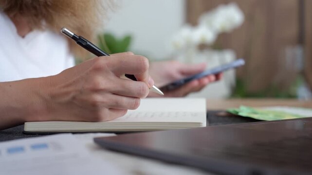 Woman planning budget writes notes in a notebook while checking her phone with euro cash on the table. Concept of household expenses and personal finance.