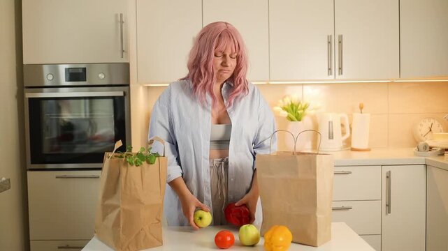 A young attractive woman with pink hair stands in a bright kitchen, holding a red bell pepper and a yellow apple while preparing healthy food.