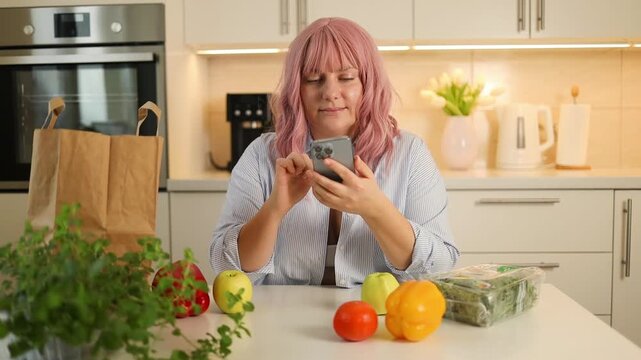 close-up shot of an attractive young woman with pink hair sitting in a bright kitchen, typing on her mobile phone near fresh vegetables and paper bags.