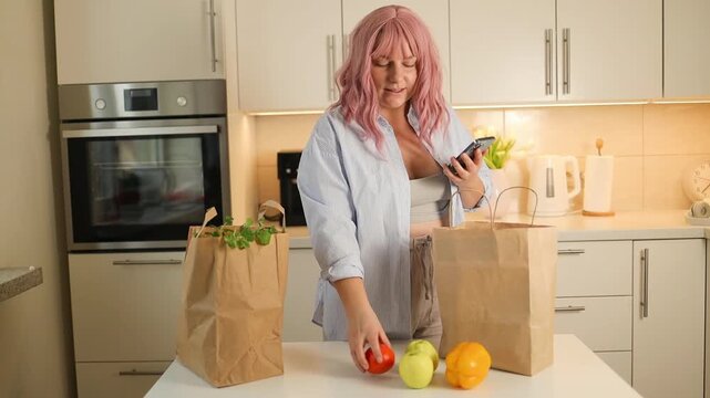 young attractive woman with pink hair stands in a bright kitchen, typing on her mobile phone next to paper shopping bags filled with fresh groceries.