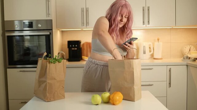 young attractive woman with pink hair stands in a bright kitchen, typing on her mobile phone next to paper shopping bags filled with fresh groceries.