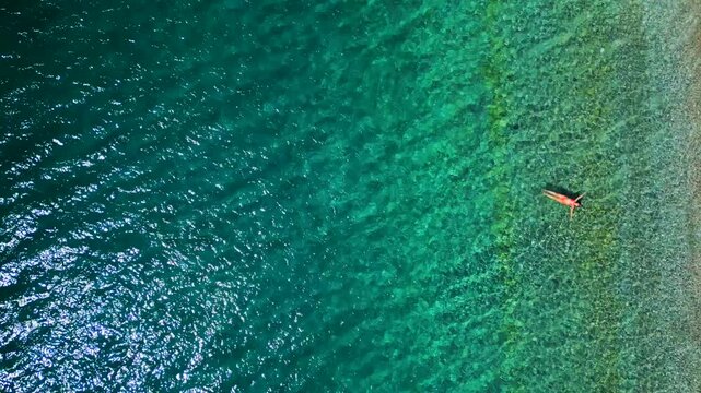 Woman in a red swimsuit lays on her back in the clear sea, top down view