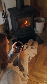Woman sitting with hot drink by the burning fireplace in the house. Winter relax