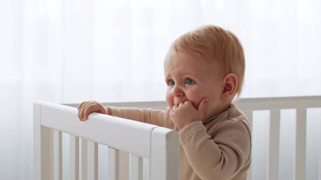 Curious Baby Boy Chewing Finger While Standing In Crib, Emitting Joyful Expression, In Bright Indoor Setting. This Adorable Infant Represents The Concept Of Baby Care And Parent-child Bonding.
