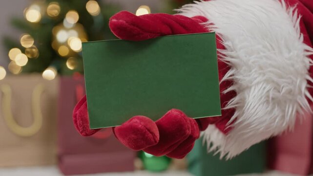 Santa Claus holding blank green card with Christmas tree and gift bags background