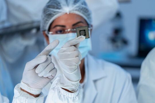 Woman in hairnet, glasses and mask holding computer processor and pointing at golden contact pins, semiconductor technology inspection.