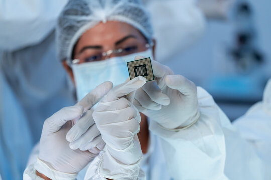 Woman in protective cap, mask and white gloves inspecting CPU microchip held in both hands, microelectronics production quality control.