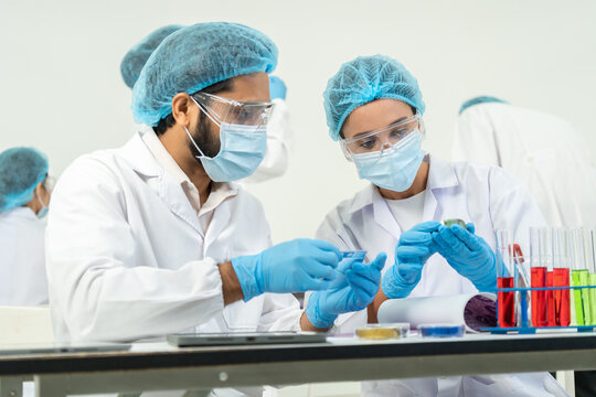 Man and woman in masks and protective caps inspecting dark rock sample, test tubes with colored liquid on bench, material analysis teamwork.