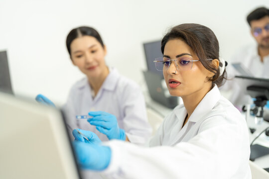 Woman in glasses inspecting sample at monitor while smiling colleague holds microscope slide, male researcher in background, bright lab.