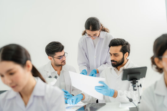 Scientists in lab coats and glasses examining document together at desk with digital microscope, collaborative data review in bright lab.