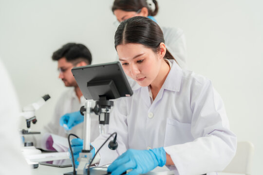 Asian woman in lab coat working with digital microscope camera at desk while colleagues work behind, modern laboratory equipment concept.