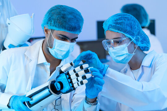 Man and woman in masks and protective caps testing bionic arm with articulated fingers, robotics and medical technology development team.