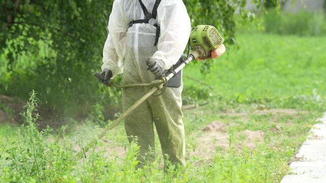 Person in white protective suit uses a handheld grass trimmer. Green grass and trees surround the garden area. Worker concentrates on trimming overgrown plants