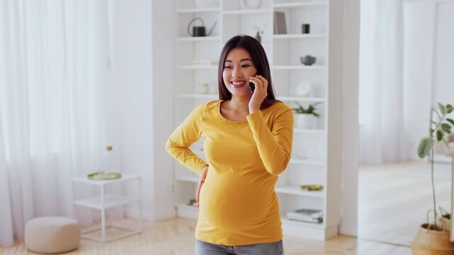 Happy Pregnant Asian Woman Talking On Smartphone While Standing In Modern Living Room, Gently Cradling Belly, Engaging In Conversation. Expecting Mother Embracing Parenthood Journey At Home.
