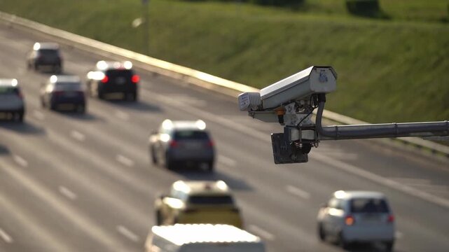 Surveillance camera mounted on a pole overlooks a highway. Cars travel on multiple lanes. Grass lines the roadside. The camera is in focus. Traffic moves in the background