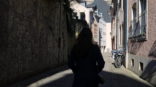 Rear view of a woman strolling through a narrow cobblestone alley flanked by tall historic stone walls