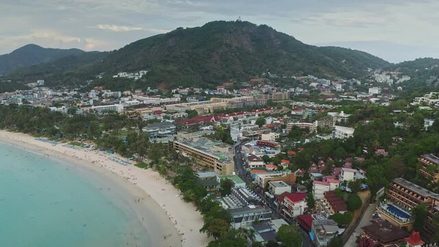 Kata Beach coastline in Phuket, Thailand, turquoise waters, sandy shore crowded with sunseekers and green hillside crowned by the Big Buddha statue, tropical resort scene. Aerial view drone flight