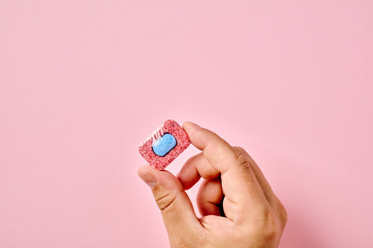 A close-up of a person's hand holding a red and blue dishwasher tablet against a pastel pink background. Minimalist flat lay composition