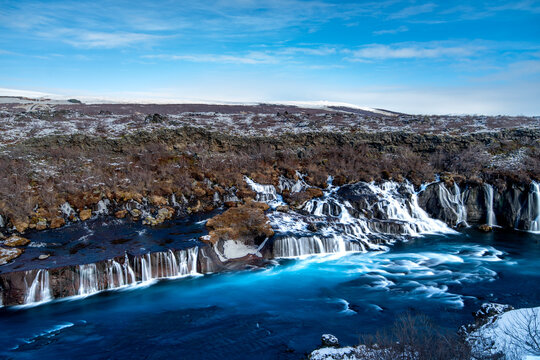 Panoramic winter view of Hraunfossar waterfalls cascading from a lava field into vibrant blue river, Iceland
