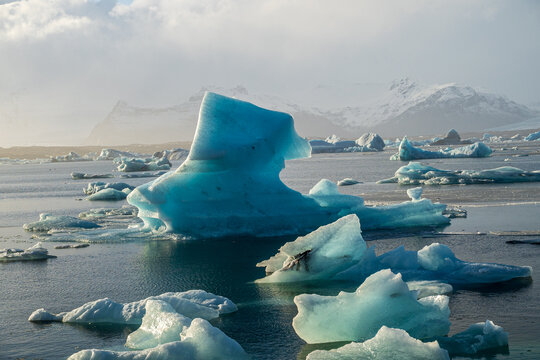 Close-up of a striking turquoise blue iceberg floating in Jokulsarlon glacier lagoon, snowy mountains in background, Iceland