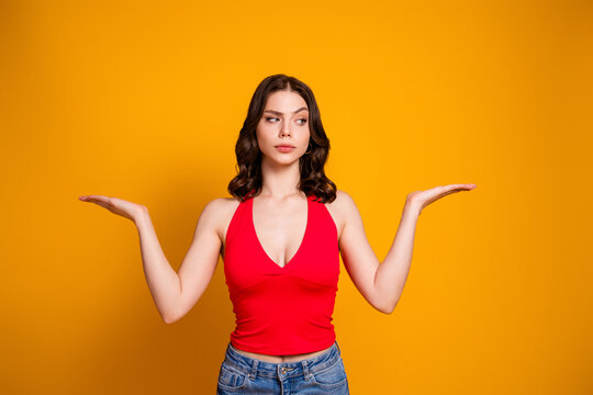 Young woman in red top weighing options with palms up against a vibrant yellow background, thoughtful expression and casual jeans conveying decision, comparison and uncertainty