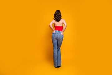 Fototapeta premium Woman standing with hands on hips wearing red top and blue jeans against vibrant orange background, rear view fashion portrait