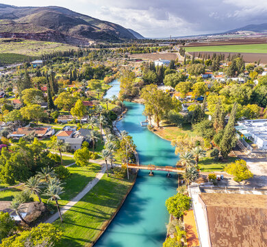 Aerial view of the Asi River flowing through the kibbutz of Nir David, surrounded by lush green trees, palm trees, and residential buildings in Nir David, North District, Israel.
