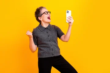 Excited woman celebrating while taking a smartphone selfie against a bright yellow background © deagreez